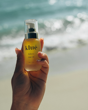Hand holding a bottle of kluē reborn hair oil by the ocean, showcasing luxury silicone-free nourishing hair oil
